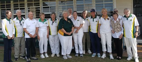 A group of eleven people in white and green croquet attire pose together outdoors in front of a building with large windows