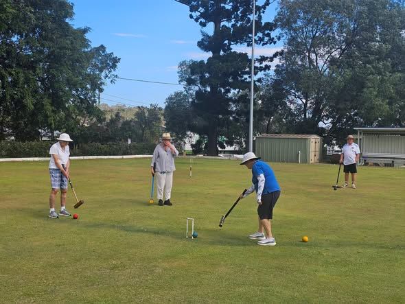 Four people play croquet on a grassy lawn, with colored balls and hoops visible under trees and a shed